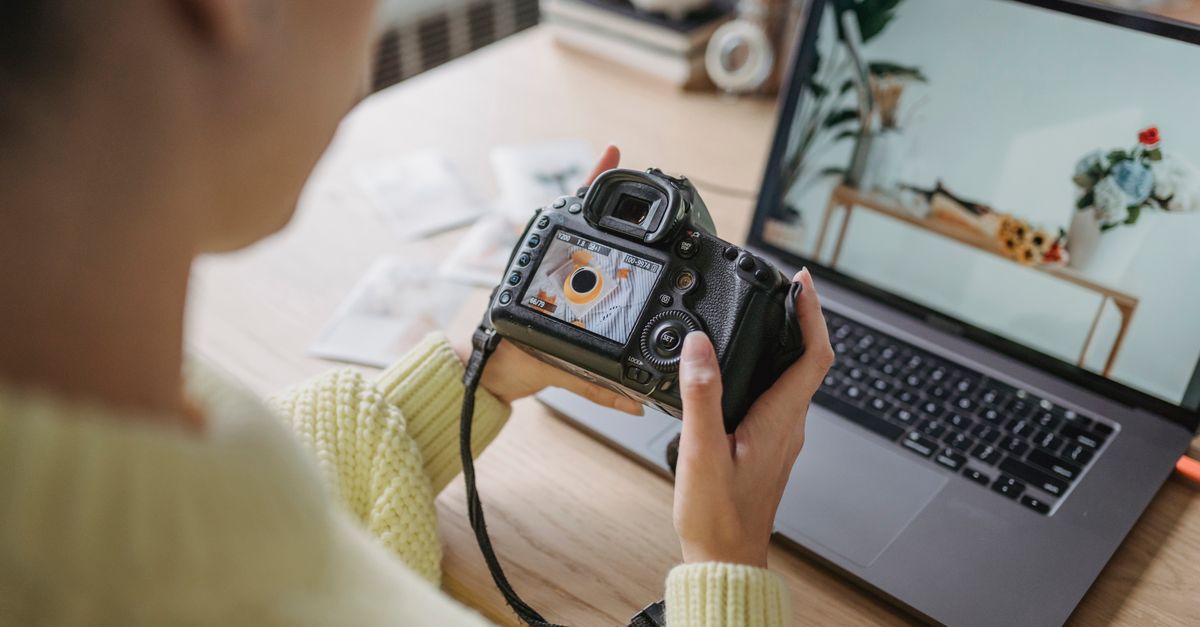 Unrecognizable crop female photographer looking at screen of photo camera while sitting at wooden table with netbook during work in light room